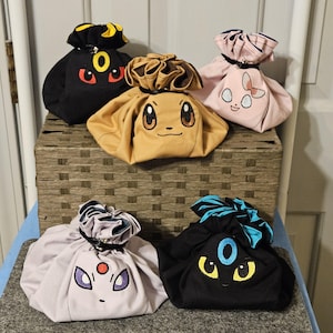 May include: Five fabric drawstring pouches featuring embroidered character faces. The collection includes a black pouch with yellow accents, a tan pouch, a light pink pouch, a light gray pouch, and a black pouch with bright blue accents.
