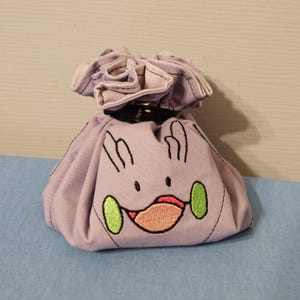 May include: A light purple drawstring pouch featuring an embroidered cartoon face with two green oval cheeks, a wide mouth with a red tongue, and two small ear-like shapes at the top. The bag is cinched at the neck with a black ribbon tie.