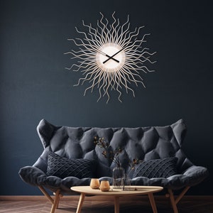 Può includere: Un orologio da parete a forma di sole con finitura in legno chiaro e lancette nere è appeso sopra un divano grigio trapuntato. Il design dell'orologio presenta linee radianti, creando un aspetto simile al sole. La stanza è decorata con un tavolino in legno, cuscini e un vaso.