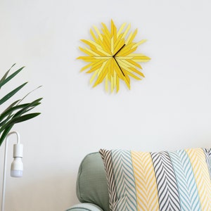 May include: A yellow and light yellow sunburst clock with black hands hanging on a white wall above a green couch with a striped pillow.