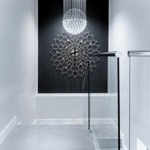 Może przedstawiać: Nowoczesny, minimalistyczny korytarz z białą ścianą i czarną ścianą, na której znajduje się duży, ozdobny zegar i kryształowy żyrandol. Szklana i metalowa balustrada prowadzi do schodów.