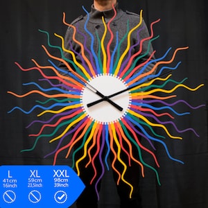 Könnte beinhalten: Eine regenbogenfarbene Uhr mit wellenförmigen Armen, die an Sonnenstrahlen erinnern. Das Zifferblatt der Uhr ist weiß mit schwarzen Zeigern. Die Uhr ist in drei Größen erhältlich: L (41 cm), XL (59 cm) und XXL (98 cm).