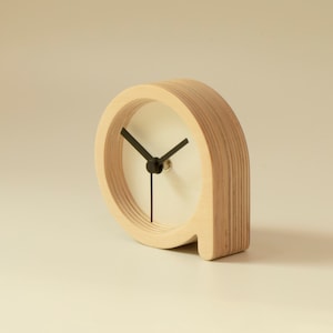 Puede incluir: Un reloj de madera con un diseño sencillo. La esfera del reloj es blanca con agujas negras y está colocada en un marco circular. El reloj tiene forma de letra 'P' y está hecho de madera en capas.