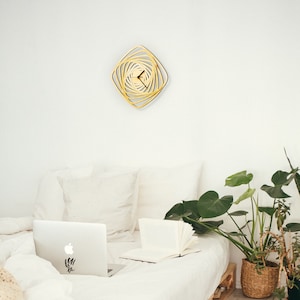 Puede incluir: Una cama blanca con ropa de cama blanca y un reloj geométrico dorado en la pared sobre la cama. Un ordenador portátil está en la cama con un libro y una planta verde en una cesta de mimbre en el suelo.