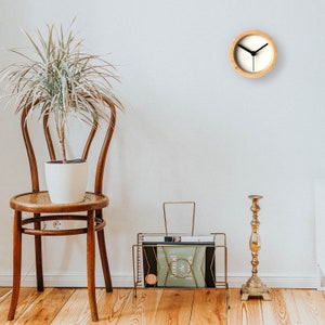 Könnte beinhalten: Eine Wanduhr aus Holz mit weißem Zifferblatt und schwarzen Zeigern. Die Uhr hängt an einer weißen Wand. Im Vordergrund sind eine Topfpflanze, ein Stuhl und ein Zeitschriftenständer zu sehen.