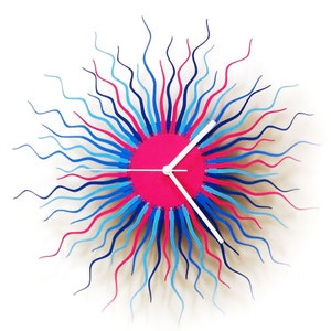 May include: A pink, blue, and white clock with wavy, sunburst-like arms. The clock face is pink with white hands.