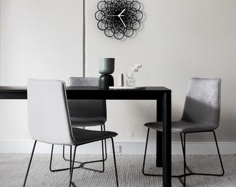 Puede incluir: Una mesa de comedor negra con cuatro sillas grises. La mesa tiene un marco de metal negro y una encimera negra. Las sillas tienen asientos de tela gris y marcos de metal negro. Un reloj negro con una esfera blanca está colgado en la pared detrás de la mesa.