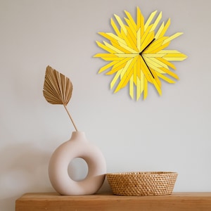 May include: A yellow sunburst clock with black hands hangs on a white wall above a beige vase with a dried palm leaf and a woven wicker basket.
