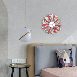 May include: A modern bedroom interior with a white and gold globe pendant light, a gray upholstered headboard, and a pink and beige wall clock with a sunburst design.