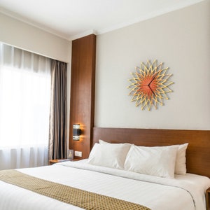 Pode incluir: Um relógio de parede decorativo com um design floral. O relógio apresenta uma moldura dourada com um centro e ponteiros vermelho-alaranjados. O relógio está montado numa parede cinza-clara, acima de uma cama com almofadas brancas e uma cabeceira castanha.