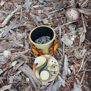 May include: A decorative, cylindrical container with a lid featuring a squirrel design, filled with sunflower seeds. The container is adorned with butterfly accents. The lid has a squirrel holding an acorn. The container is on a bed of wood chips.
