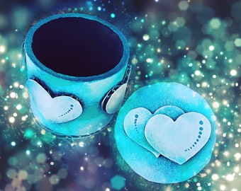Può includere: Una scatola cilindrica turchese con coperchio abbinato. La scatola e il coperchio sono adornati con decorazioni bianche a forma di cuore. La scatola è aperta, rivelando un interno scuro. Lo sfondo è un bokeh di luci turchesi e bianche.