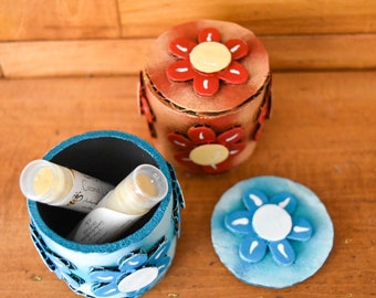May include: Three handmade, decorative containers with floral designs. One is a cylindrical, light blue container with a lid and blue flower accents. Another is a brown container with red flower accents. The third is a brown container with a lid and red flower accents.