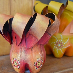 Peut inclure: Deux pots de fleurs décoratifs en plastique recyclé. Les pots sont peints dans des tons de rouge, orange et jaune avec des pois blancs et des motifs floraux.