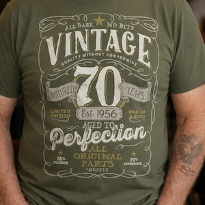 May include: An olive green t-shirt featuring a graphic design with the text VINTAGE 70 YEARS AGED TO PERFECTION EST 1956. The print includes decorative flourishes, stars, and additional text reading ALL BARK NO BITE, QUALITY WITHOUT COMPROMISE, and ALL ORIGINAL PARTS.