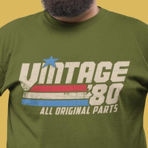 May include: A green t-shirt with a vintage-style graphic that says "Vintage '80 All Original Parts". The graphic features a red, white, and blue stripe design with a star.