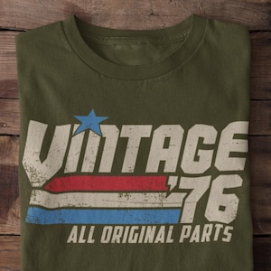 May include: Olive green t-shirt with a distressed "VINTAGE '76" graphic. The design includes a blue star, red and blue stripes, and the text "ALL ORIGINAL PARTS". The shirt is folded on a wooden surface.