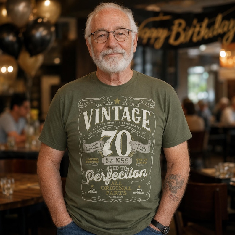 May include: An olive green t-shirt featuring a graphic design with the text VINTAGE 70 YEARS AGED TO PERFECTION EST 1956. The print includes decorative flourishes, stars, and additional text reading ALL BARK NO BITE, QUALITY WITHOUT COMPROMISE, and ALL ORIGINAL PARTS.