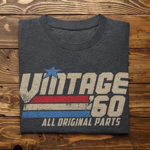 May include: Dark gray t-shirt with the text "VINTAGE '60 ALL ORIGINAL PARTS". The design includes a blue star, red and white stripes, and blue stripes. The shirt is folded on a wooden surface.