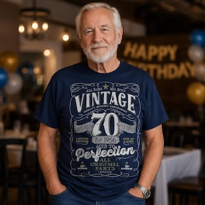 May include: A navy blue t-shirt featuring a white graphic design with the text Vintage 70 Years Matured Aged to Perfection Est 1956 All Original Parts. The print includes decorative scrollwork, stars, and various phrases in a vintage-style layout.