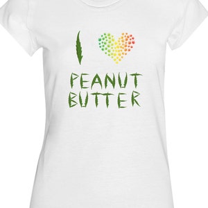 Può includere: Maglietta bianca con una foglia di marijuana verde e un cuore fatto di foglie di marijuana verdi in colori arcobaleno. Il testo "Amo il burro di arachidi" è scritto in verde sotto il cuore.