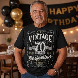 May include: A black t-shirt featuring a white graphic design with the text "VINTAGE 70 YEARS MATURED EST 1956 AGED TO PERFECTION ALL ORIGINAL PARTS". The design includes decorative scrollwork, stars, and various phrases in a vintage-inspired layout.