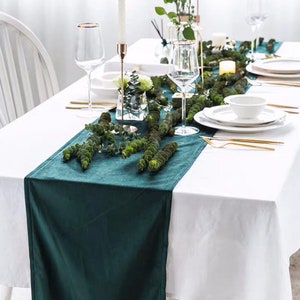 Op de afbeelding: Een formele eettafel met een wit tafelkleed en een donkergroene tafelloper. De tafel is gedekt met witte borden, gouden bestek en heldere wijnglazen. Bloemschikkingen en kaarsen completeren de elegante inrichting.