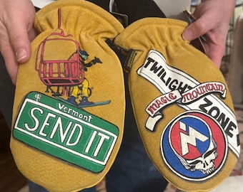 May include: Mustard yellow ski mittens with graphic designs. One mitten features a skier on a ski lift and a Vermont "SEND IT" street sign. The other has "TWILIGHT MOUNTAIN ZONE" text and a skull logo.