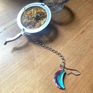 May include: A stainless steel tea infuser with a chain and a decorative crescent moon charm. The infuser is filled with dried herbs and sits on a wooden surface. The charm has a gradient of purple and teal.