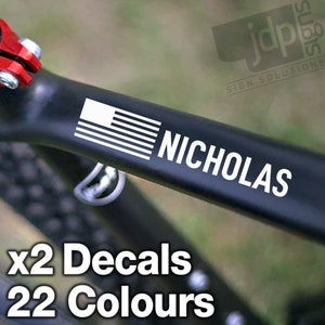May include: Two black and white decals with an American flag and the name "NICHOLAS" in white text. The decals are on a black bicycle frame. The text "x2 Decals 22 Colours" is at the bottom of the image.