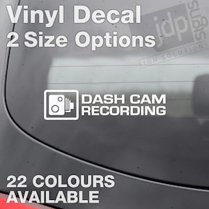 Op de afbeelding: Witte vinyl sticker met de tekst "DASH CAM RECORDING" en een camera-icoon. De sticker is verkrijgbaar in 22 kleuren en twee maten.