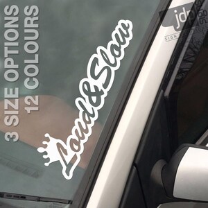 Op de afbeelding: Witte vinyl sticker met de woorden "Loud & Slow" in een cursief lettertype, met een kroonafbeelding. De sticker zit op een autoruit. De tekst "3 SIZE OPTIONS 12 COLOURS" is ook zichtbaar.