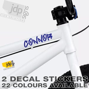 May include: Two blue decal stickers with the name "CONNOR" in a graffiti style font. The stickers are shown on a white bicycle frame. The text "2 DECAL STICKERS 22 COLOURS AVAILABLE" is also shown in the image.