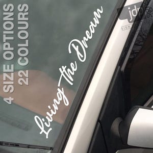 May include: White vinyl decal sticker with the text "Living the Dream" on a car window. The sticker is on the driver's side window and is angled diagonally.