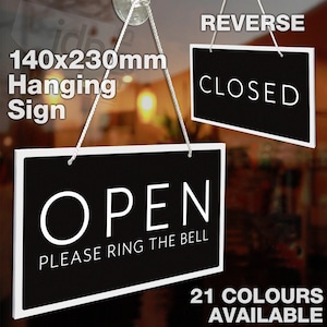 May include: A black and white hanging sign with the word "OPEN" in white letters and the text "PLEASE RING THE BELL" below. The sign is 140x230mm and has a white border. The sign is hanging from a string and is in front of a blurry background. The sign also has a reverse side that says "CLOSED" in white letters on a black background.