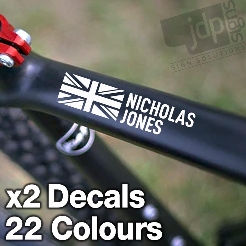 K&ouml;nnte beinhalten: Zwei wei&szlig;e Vinyl-Aufkleber mit einer Union Jack-Flagge und dem Namen "Nicholas Jones" in schwarz-wei&szlig;er Schrift. Die Aufkleber sind auf einem schwarzen Fahrradrahmen angebracht. Der Text "x2 Decals 22 Colours" ist ebenfalls auf dem Bild zu sehen.
