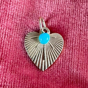 May include: A heart-shaped metal pendant featuring a sunburst pattern of engraved lines radiating from the centre. A small, round, bright turquoise-coloured stone is set at the top of the heart, just below the attached silver-toned jump ring.