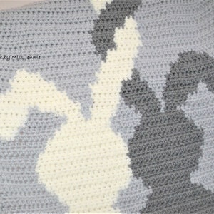 May include: A crocheted baby blanket with a gray, white, and cream color scheme. The blanket features a pattern of bunnies in a camouflage style. The text "Handmade by Miss Jennie" is visible in the top left corner.