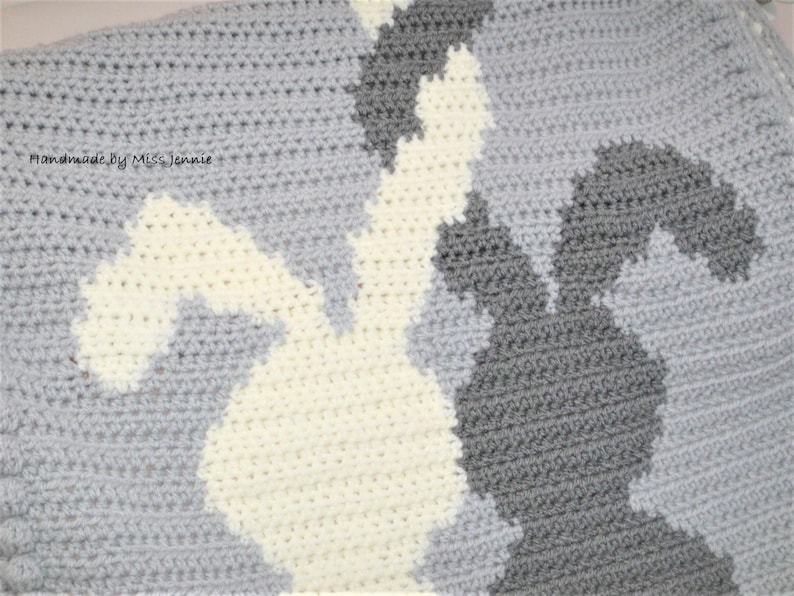 May include: A crocheted baby blanket with a gray, white, and cream color scheme. The blanket features a pattern of bunnies in a camouflage style. The text "Handmade by Miss Jennie" is visible in the top left corner.