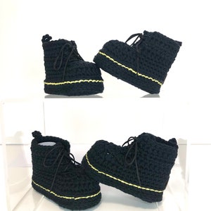 May include: A pair of black crocheted baby booties with yellow stitching. The booties have a lace-up design and a chunky platform sole.