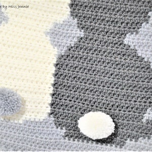 May include: A crocheted baby blanket with a yin yang design in white and gray. The blanket has two pom-poms, one white and one gray.