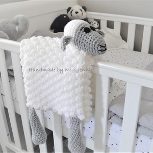 May include: White and grey crocheted baby blanket with a sheep head and legs. The blanket is hanging on a white cot with a black and white star patterned sheet. The text "Handmade by Miss Jennie" is visible on the blanket.