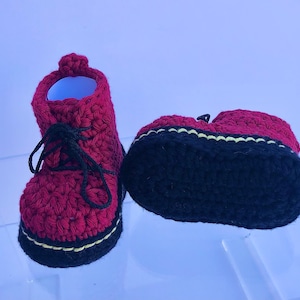 May include: A pair of red crocheted baby booties with black laces and a black and yellow trim. The booties have a black crocheted sole.