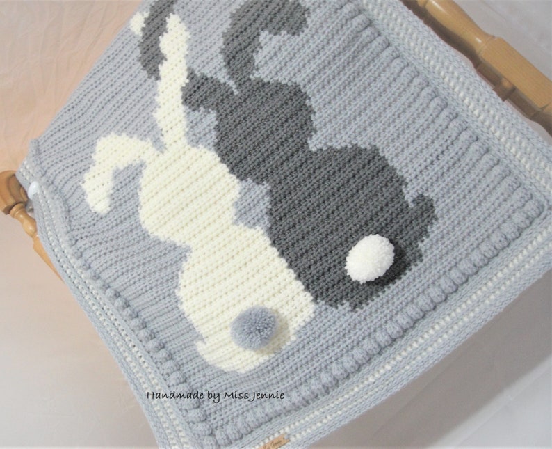 May include: A gray and white crocheted baby blanket with a design of two bunnies. The bunnies have white pom-poms for tails. The blanket has a gray border with a white trim. "Handmade by Miss Jennie" is written on the blanket.