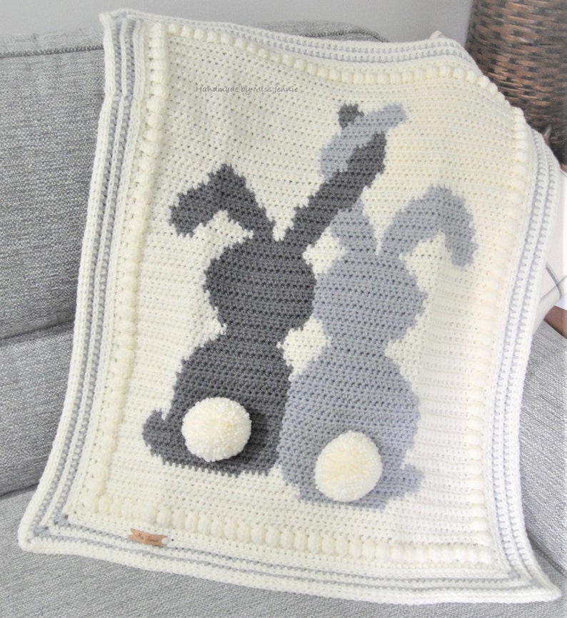 May include: A crocheted baby blanket with two gray bunny silhouettes and white pom-poms. The blanket has a white background with a white and gray border.