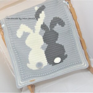 May include: Handmade by Miss Jennie, a gray crocheted baby blanket with two white and gray bunny silhouettes. The bunnies have white pom-poms for tails.