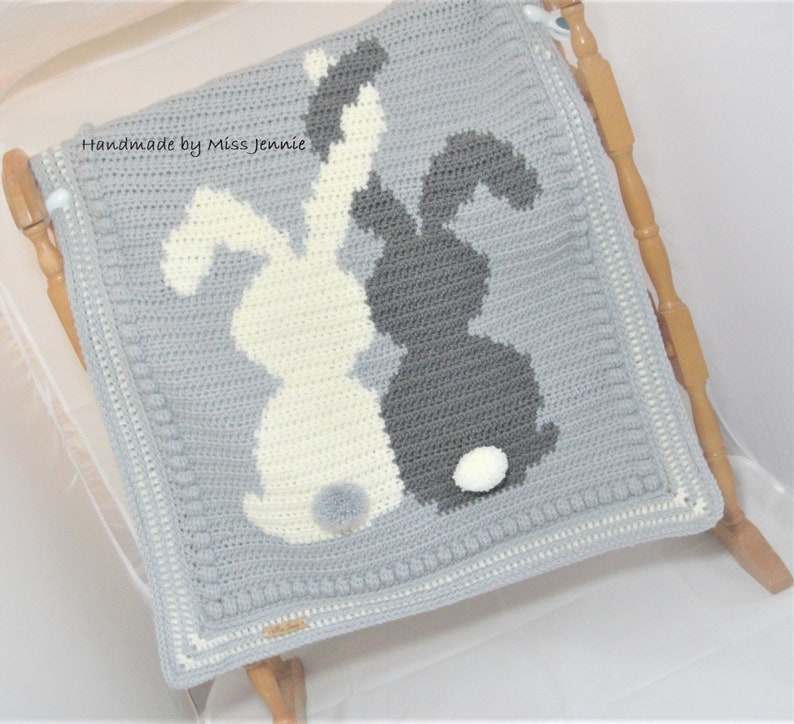May include: Handmade by Miss Jennie, a gray crocheted baby blanket with two white and gray bunny silhouettes. The bunnies have white pom-poms for tails.