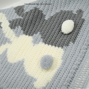May include: A gray and white crocheted baby blanket with two bunny shapes. The bunnies have white pom-poms for tails.
