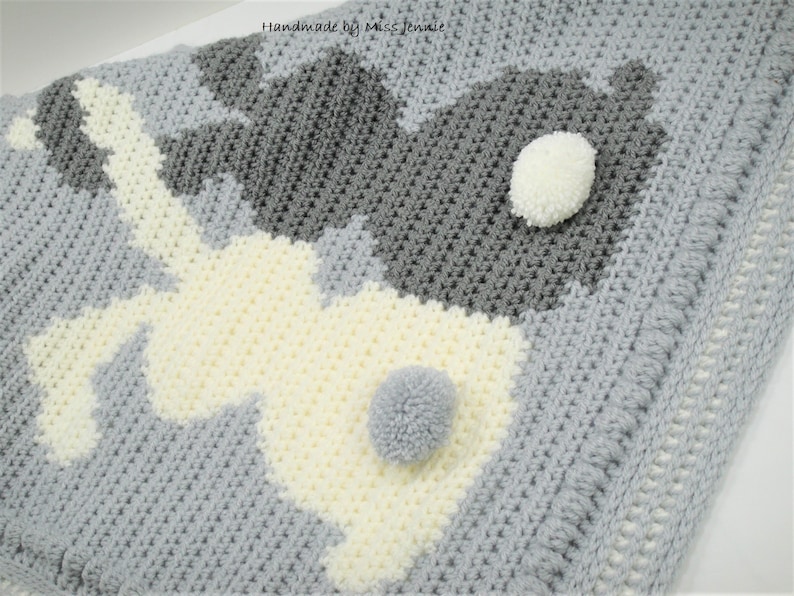 May include: A gray and white crocheted baby blanket with two bunny shapes. The bunnies have white pom-poms for tails.