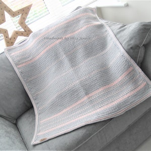 May include: A gray and pink knitted blanket with horizontal stripes, resting on a gray sofa. The blanket has a textured appearance and a light pink border. The text "Handmade by miss Jennie" is visible on the blanket. A wooden star decoration is in the background.